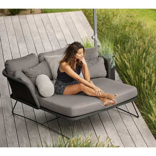 lifestyle image of cane-line horizon daybed in Black, Small Round Cane-line Weave Incl. Grey Cane-line Natté in a patio with a woman enjoying the daybed