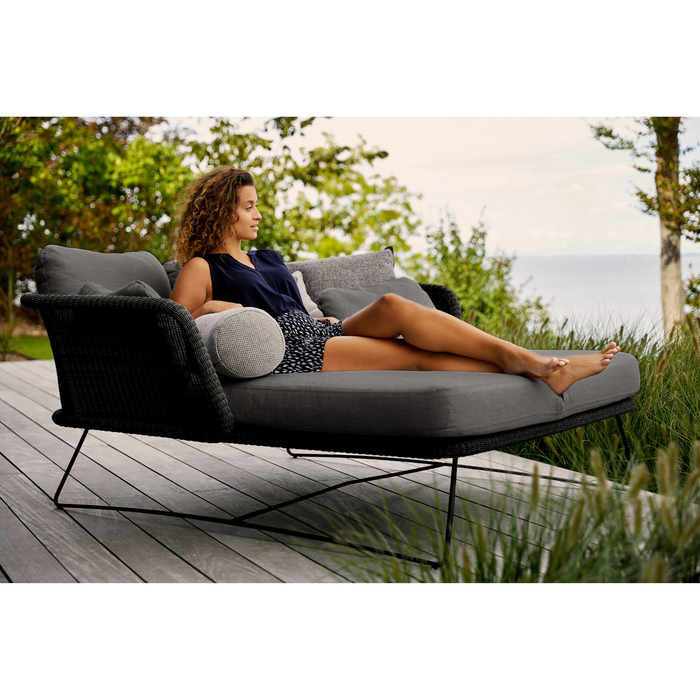 lifestyle image of cane-line horizon daybed in Black, Small Round Cane-line Weave Incl. Grey Cane-line Natté  with parasol in a patio and a woman enjoying the daybed