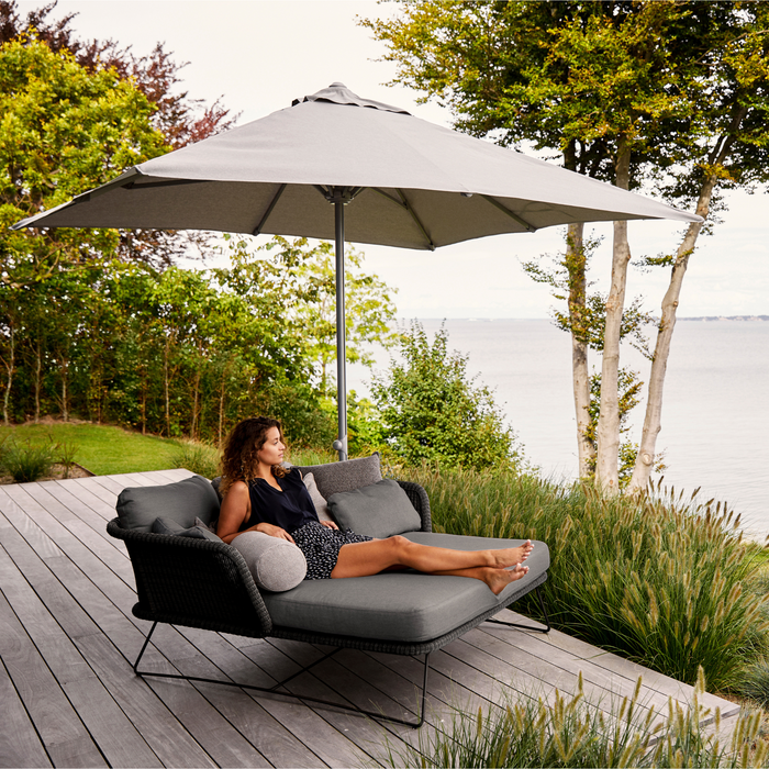 lifestyle image of cane-line horizon daybed in Black, Small Round Cane-line Weave Incl. Grey Cane-line Natté in a patio with a woman enjoying the daybed