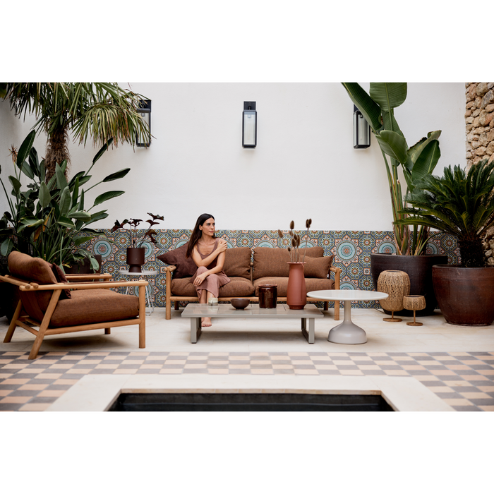 sticks 2 seater sofa Teak sofa Umber brown Cane-line Rise cushion in a patio with outdoor furniture and a woman enjoying the sofa lifestyle