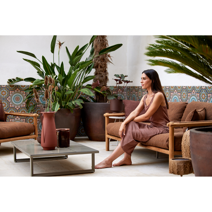 sticks 2 seater sofa Teak sofa Umber brown Cane-line Rise cushion in a patio with outdoor furniture and a woman enjoying the sofa lifestyle