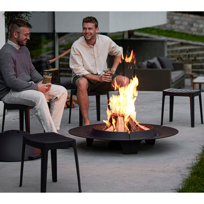 lifestyle image of cane-line cut stool in Black, Aluminium chair without cushion in a patio around a fire pit with two men enjoying the chair