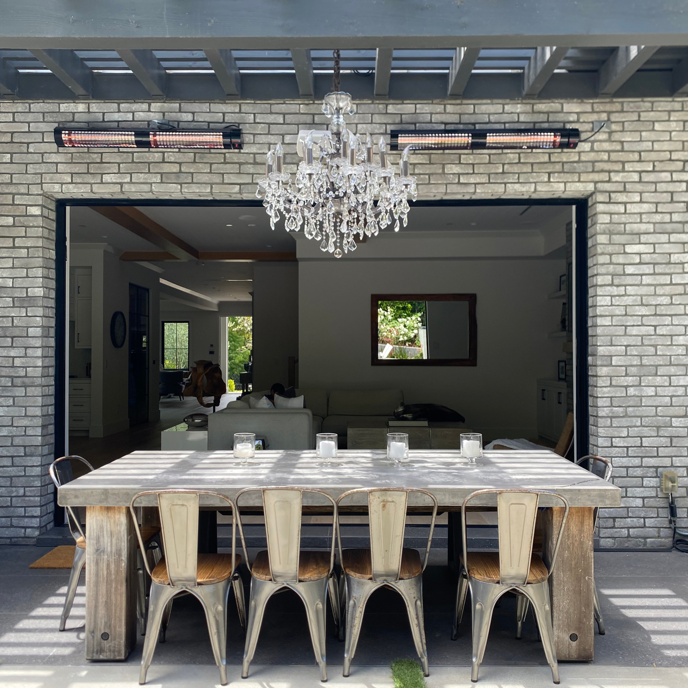 Dining area with a large wooden table and chairs in front of a brick wall with a chandelier with the heaters mounted to the brick walls.