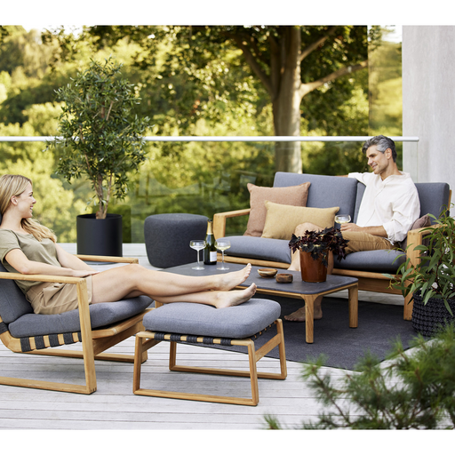 lifestyle image of cane-line endless soft footstool in Dark Grey, Medium Flat Cane-line Soft Rope w/Teak Incl. Grey Cane-line Natté in a patio with outdoor furniture and a woman enjoying a chair and the footstool