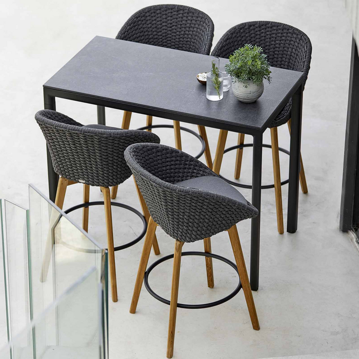 lifestyle image of cane-line peacock bar chair in Dark Grey, Medium Flat Cane-line Soft Rope w/Teak chair with Grey, Cane-line Natté cushion in a patio with bar table