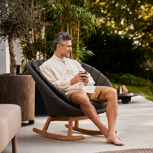 cane-line peacock rocking chair Dark Grey, Medium Flat Cane-line Soft Rope with Teak and Dark Grey, Cane-line Focus cushion in a patio with a man enjoying the chair lifestyle