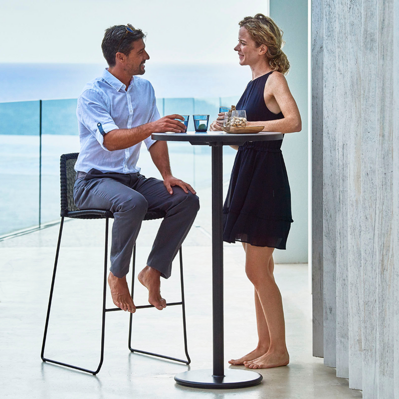 lifestyle image of cane-line breeze bar chair in Black, Small Round Cane-line Weave with bar table in a patio and a man enjoying the chair