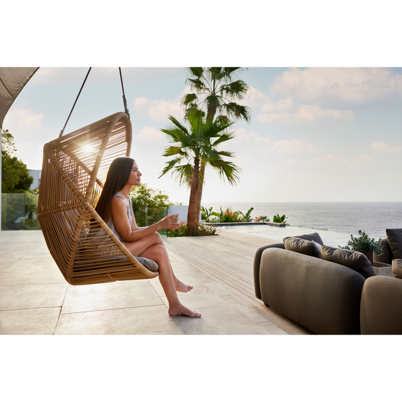 cane line hive chair Natural, small round tube Cane-line Weave and Taupe, string Cane-line Soft Rope and Taupe, Cane-line AirTouch cushion in a patio with a woman enjoying the chair lifestyle