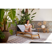 cane-line siesta sunbed teak sunbed with Light grey, Cane-line Virtue cushion in a patio with plants and a woman enjoying the sunbed lifestyle