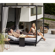 lifestyle image of cane-line laze cabana in Lava Grey, Aluminium Incl. Light Grey Cane-line Natté in a patio with outdoor furniture and two people enjoying the cabana