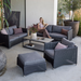 lifestyle image of cane-line diamond footstool in Graphite, Small Round Cane-line Weave Incl. Grey Cane-line Natté with outdoor furniture in a patio and a woman enjoying a sofa